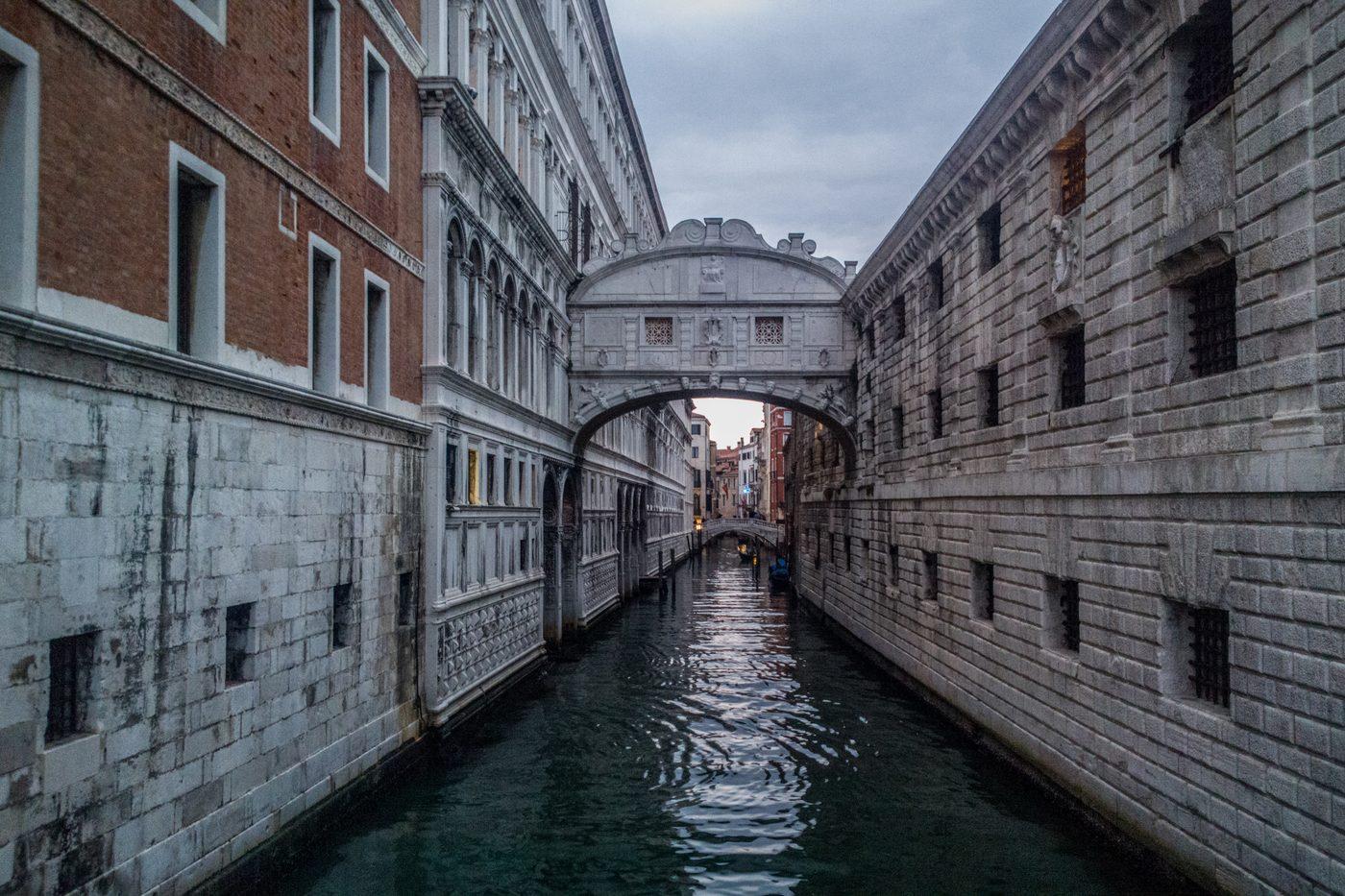 A narrow Venetian canal running beneath the Bridge of Sighs at dusk, empty of boats
